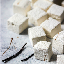 Load image into Gallery viewer, Tabletop with white puffy square marshmallows dusted with powdered sugar, and drizzled with vanilla, decorated with two whole vanilla beans..