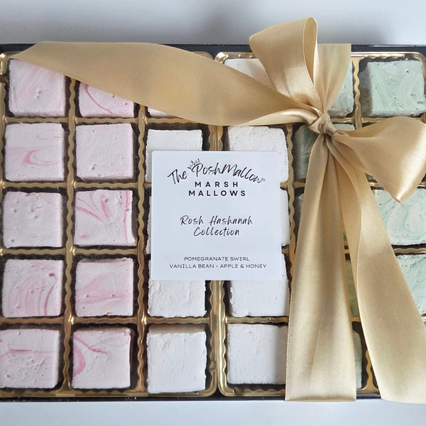 A boxed assortment of marshmallows from The PoshMallow, featuring a black tray with gold trim, pale pink and mint green colored marshmallows, a white paper label, and a large gold ribbon tied in a bow on a light gray background.