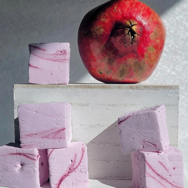 Four pink cube marshmallows with pink marble veining are arranged on white pedestals beneath a red pomegranate, photographed in strong sunlight against a gray wall.