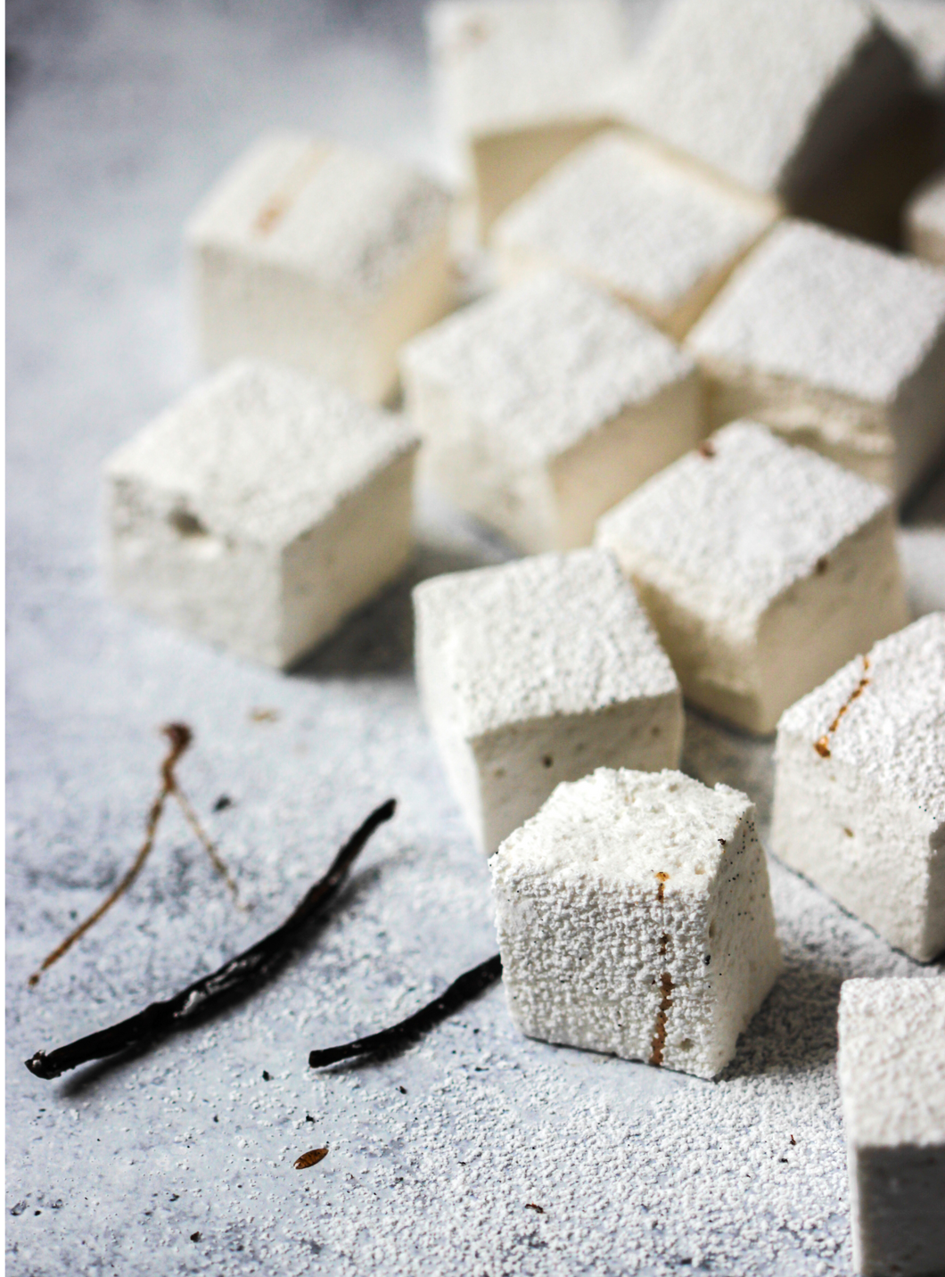 Tabletop with 12 white puffy square marshmallows dusted with powdered sugar, and drizzled with vanilla, decorated with two whole vanilla beans..